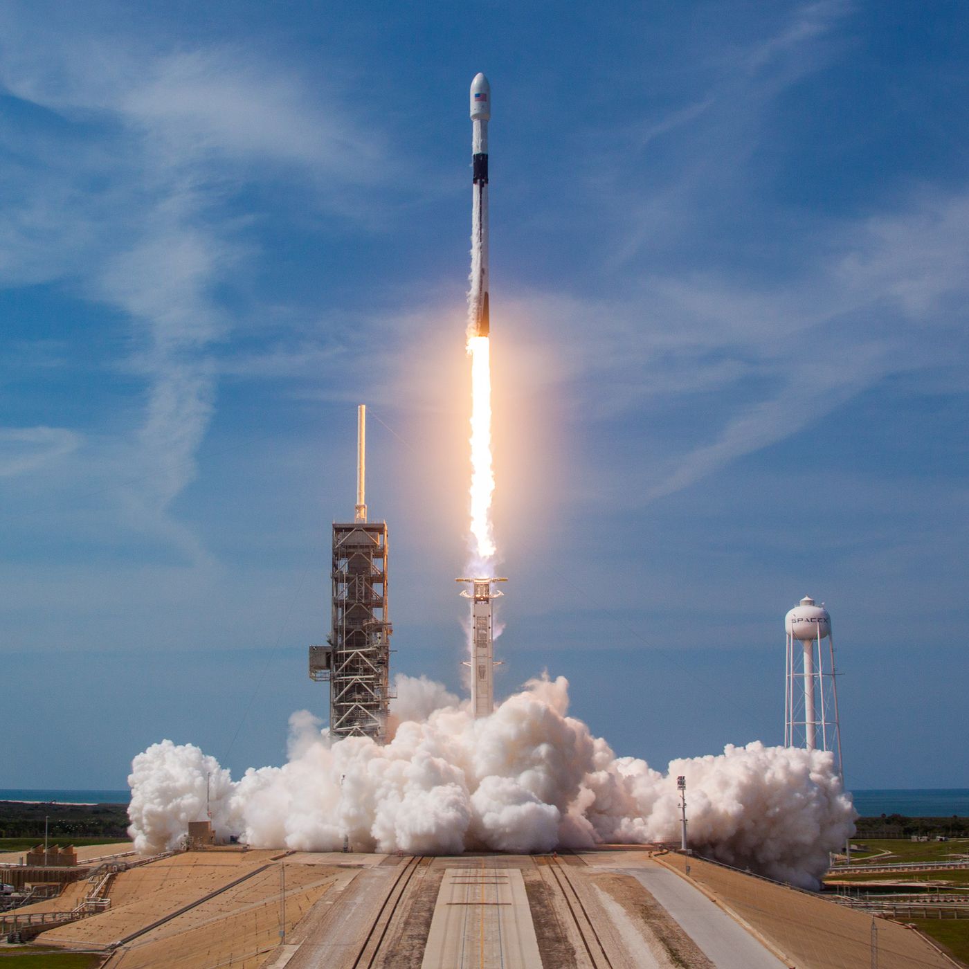 Falcon 9 launching from its launch pad. Cloud of smoke covers the base of the pad which looks like a large concrete runway. The sky is very blue with some fine white clouds.